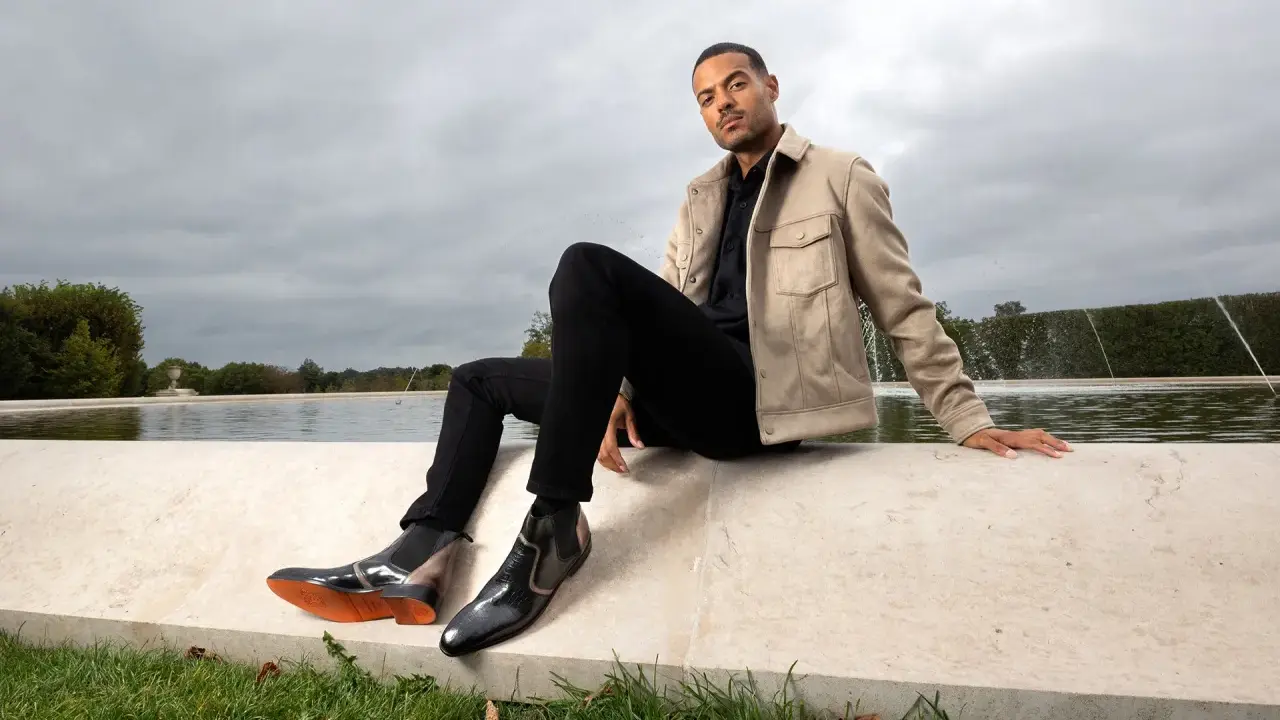 Mann in schwarzer Hose, beigem Sakko und stylischen Herren Boots, sitzt am Wasser.