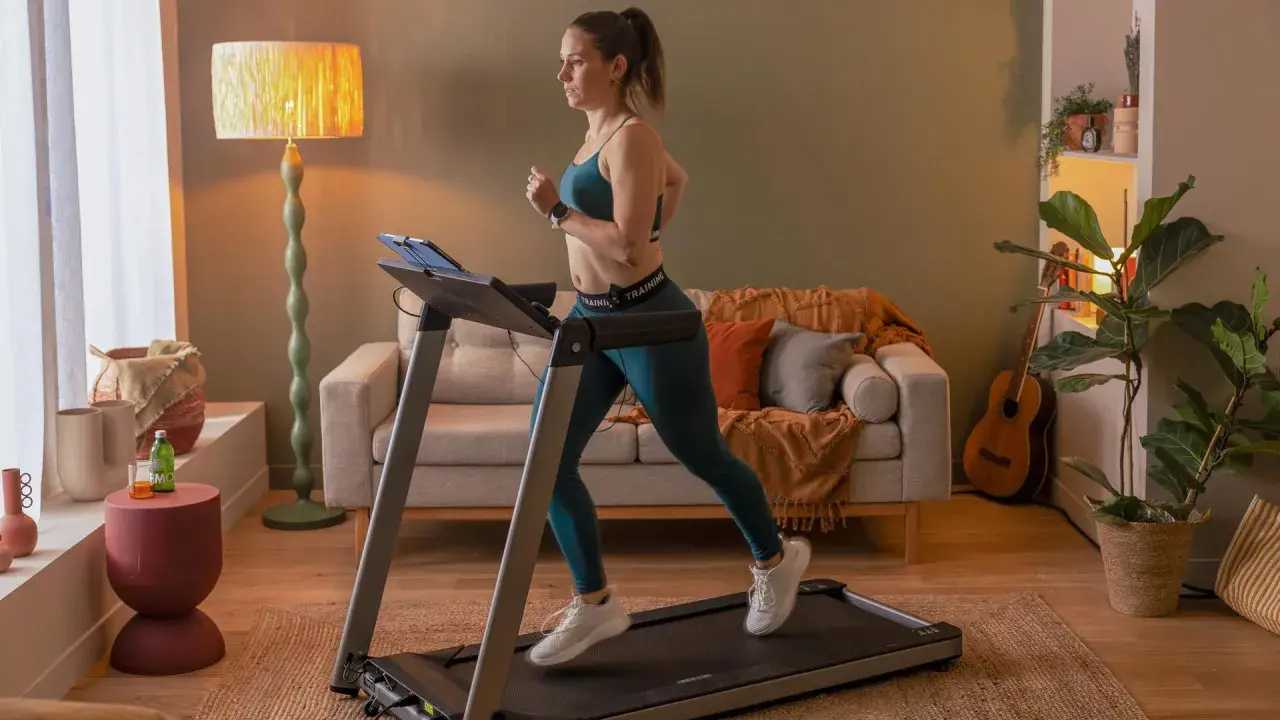 Frau macht Intervalltraining auf Laufband im Wohnzimmer. Sie trägt Sportkleidung und läuft mit Energie.