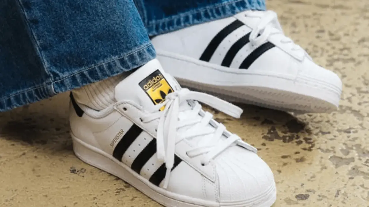 Weiße Adidas Superstar Sneaker mit schwarzen Streifen und gelbem Logo. Perfekt, um das Adidas modell herausfinden.