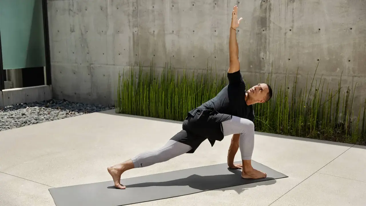 Mann in Yoga-Pose auf Matte vor Betonwand und grünen Pflanzen.
