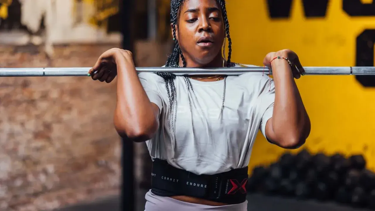 Frau mit geflochtenen Haaren hebt eine Hantel für Jumping Squats. Sie trägt einen Trainingsgürtel.