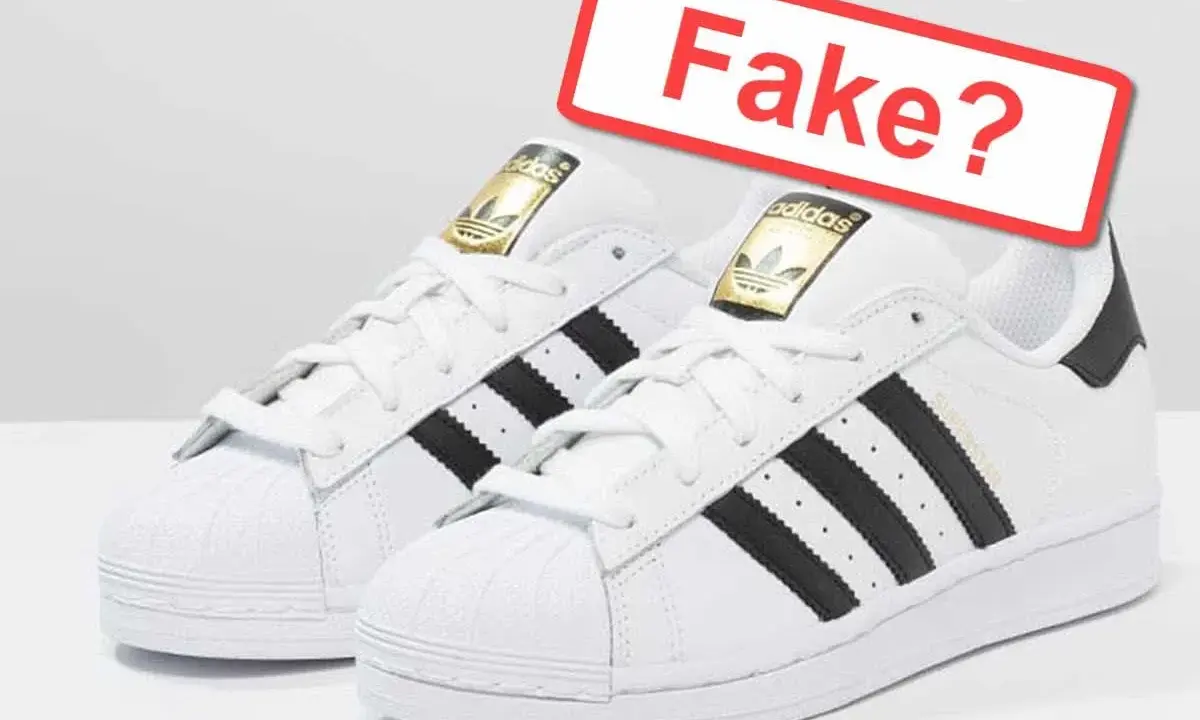 Zwei weiße Adidas Superstar Sneaker mit schwarzen Streifen. Ein rotes Schild fragt: "Fake?". So kannst du Adidas fake erkennen.