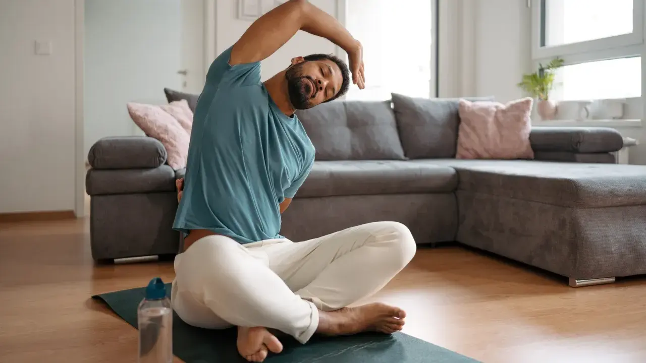 Mann macht Dehnen am Morgen auf einer Matte im Wohnzimmer. Er sitzt im Schneidersitz und beugt sich zur Seite.