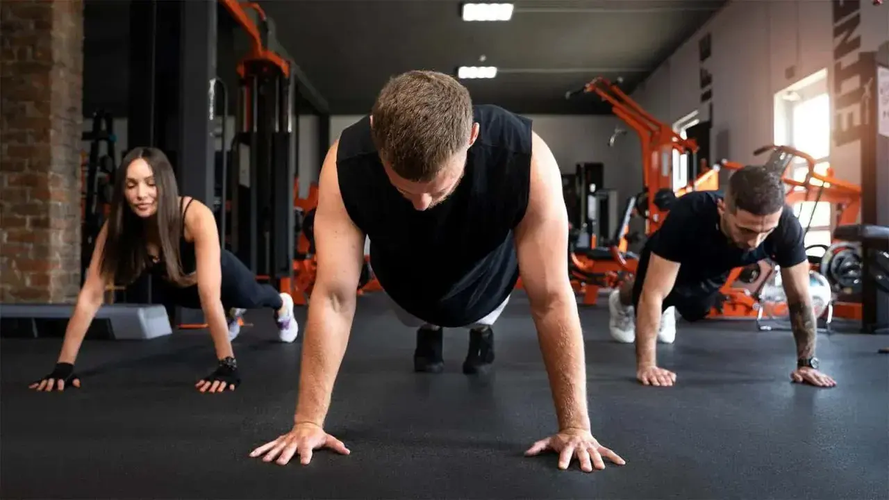 Drei Personen machen Liegestütze im Fitnessstudio. Sie lernen, was sind Burpees, eine Ganzkörperübung.
