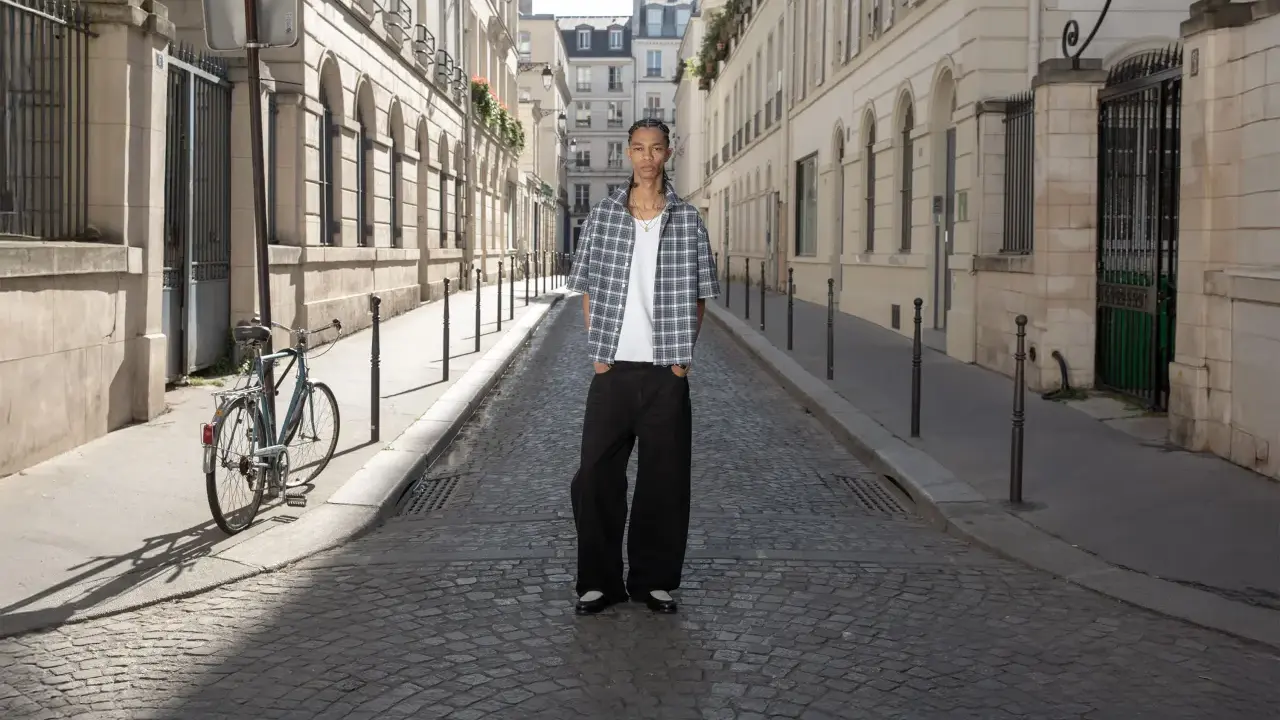 Ein Mann steht auf einer Kopfsteinpflasterstraße in Paris. Er trägt ein kariertes Hemd und weite Hosen. Ein Fahrrad lehnt an einem Laternenpfahl. Das Bild strahlt eine zeitlose Eleganz aus, die an reternity deutsch erinnert.