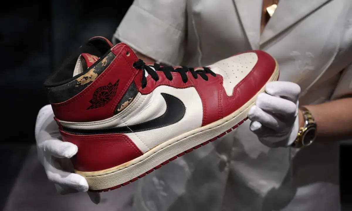 Ein Paar abgenutzte Air Jordan 1 Sneaker, die seit 1985 erhältlich sind, werden von einer Person mit weißen Handschuhen gehalten.