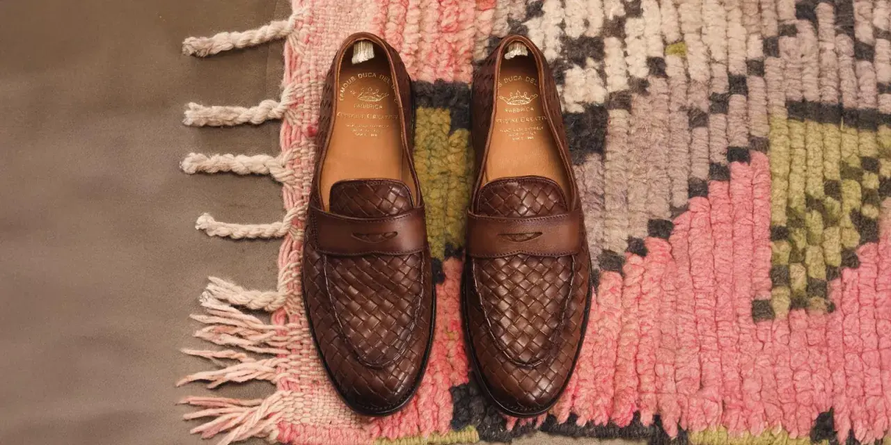 Zwei braune Leder-Loafer mit geflochtenem Muster, Marke Officine Creative, liegen auf einem bunten Teppich.