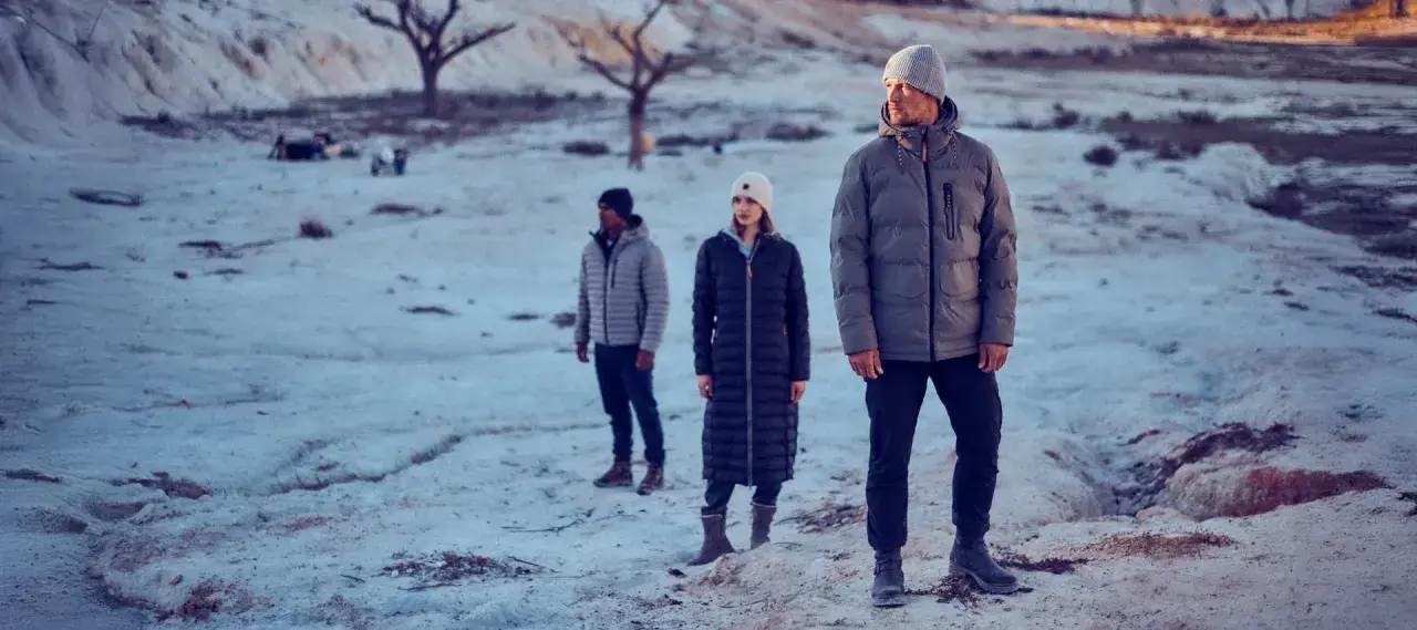 Drei Personen in Winterjacken stehen in einer verschneiten, kargen Landschaft. Die Frage "ab wann Winterjacke" stellt sich bei diesem Anblick von selbst.