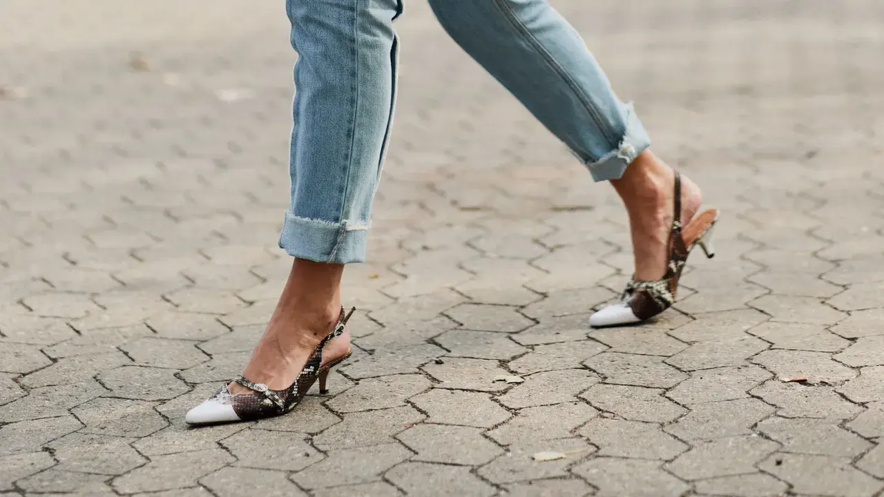 Beine in hochgekrempelten Jeans und Schlangenleder-Pumps auf gepflastertem Untergrund.