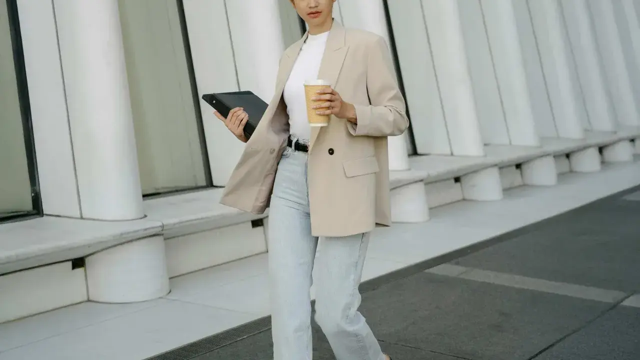 Frau in **smart casual** Kleidung mit Blazer, Jeans und Kaffeebecher geht mit Tablet.
