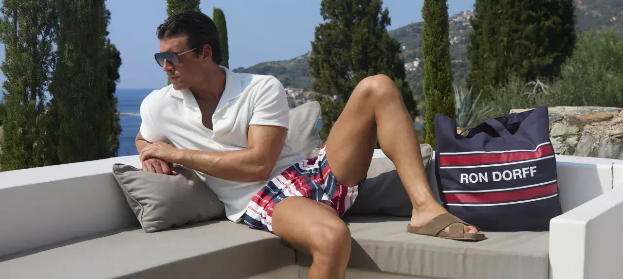 Ein Mann mit Sonnenbrille und gestreifter Badehose entspannt sich auf einer Couch. Neben ihm liegt eine Tasche mit der Aufschrift "RON DORFF".
