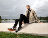 Mann in schwarzer Hose, beigem Sakko und stylischen Herren Boots, sitzt am Wasser.