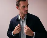Ein Mann mit Bart und kurz geschnittenem Haar, der eine dunkelblaue Jacke über einem gestreiften Hemd trägt. Er fasst nach dem Reißverschluss, als würde er seine Kleidung für das Hemden waschen vorbereiten.
