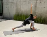 Mann in Yoga-Pose auf Matte vor Betonwand und grünen Pflanzen.