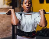 Frau mit geflochtenen Haaren hebt eine Hantel für Jumping Squats. Sie trägt einen Trainingsgürtel.