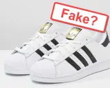 Zwei weiße Adidas Superstar Sneaker mit schwarzen Streifen. Ein rotes Schild fragt: "Fake?". So kannst du Adidas fake erkennen.
