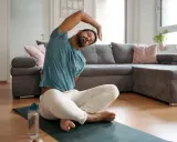 Mann macht Dehnen am Morgen auf einer Matte im Wohnzimmer. Er sitzt im Schneidersitz und beugt sich zur Seite.