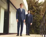 Zwei Männer in eleganten Anzügen, die aktuelle Anzug Trends verkörpern, stehen auf einer Holzterrasse vor einem modernen Gebäude.