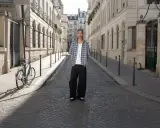 Ein Mann steht auf einer Kopfsteinpflasterstraße in Paris. Er trägt ein kariertes Hemd und weite Hosen. Ein Fahrrad lehnt an einem Laternenpfahl. Das Bild strahlt eine zeitlose Eleganz aus, die an reternity deutsch erinnert.