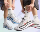 Zwei Paare Nike P-6000 Sneaker in Weiß mit blauen und roten Akzenten. Die Füße tragen weiße Socken mit Nike-Logo und auffälligen Schmuck.