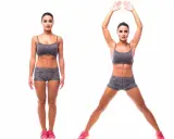 Eine Frau zeigt, was sind Jumping Jacks: erst mit geschlossenen Beinen, dann mit gespreizten Beinen und erhobenen Armen.