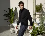 Mann in schwarzer Jacke und blauer Hose lehnt an Säule, im Hintergrund Pflanzen und Tische. Gran Sasso Mode.