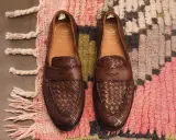 Zwei braune Leder-Loafer mit geflochtenem Muster, Marke Officine Creative, liegen auf einem bunten Teppich.