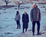 Drei Personen in Winterjacken stehen in einer verschneiten, kargen Landschaft. Die Frage "ab wann Winterjacke" stellt sich bei diesem Anblick von selbst.