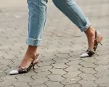 Beine in hochgekrempelten Jeans und Schlangenleder-Pumps auf gepflastertem Untergrund.
