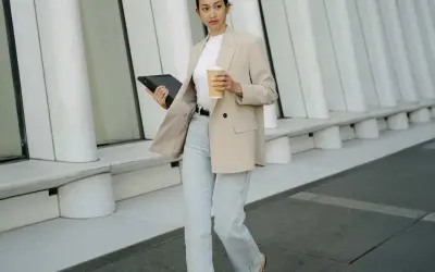 Frau in **smart casual** Kleidung mit Blazer, Jeans und Kaffeebecher geht mit Tablet.