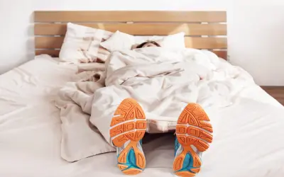 Füße mit Laufschuhen ragen aus einer Decke. Der Wecker klingelt, aber die Person träumt noch vom morgens joggen.