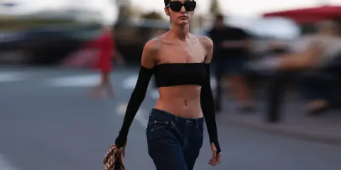 Frau in typischer 2000er Mode: schwarzer Bandeau-Top, tief sitzende Jeans und Armstulpen. Sie trägt Sonnenbrille und eine Tasche.