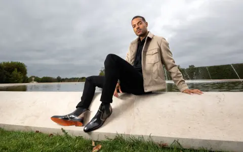 Mann in schwarzer Hose, beigem Sakko und stylischen Herren Boots, sitzt am Wasser.