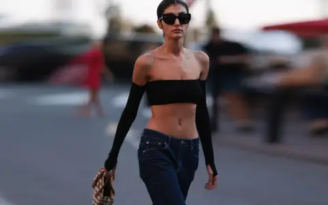 Frau in typischer 2000er Mode: schwarzer Bandeau-Top, tief sitzende Jeans und Armstulpen. Sie trägt Sonnenbrille und eine Tasche.