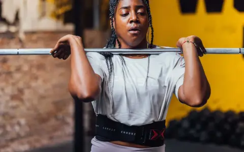 Frau mit geflochtenen Haaren hebt eine Hantel für Jumping Squats. Sie trägt einen Trainingsgürtel.