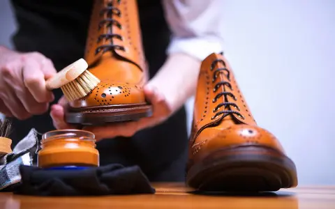 Hände bürsten braune Lederschuhe, um sie zu reinigen. Daneben steht eine Dose Schuhcreme.