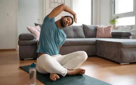 Mann macht Dehnen am Morgen auf einer Matte im Wohnzimmer. Er sitzt im Schneidersitz und beugt sich zur Seite.