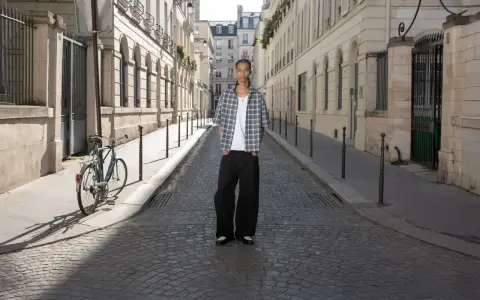 Ein Mann steht auf einer Kopfsteinpflasterstraße in Paris. Er trägt ein kariertes Hemd und weite Hosen. Ein Fahrrad lehnt an einem Laternenpfahl. Das Bild strahlt eine zeitlose Eleganz aus, die an reternity deutsch erinnert.