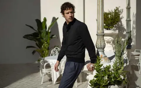 Mann in schwarzer Jacke und blauer Hose lehnt an Säule, im Hintergrund Pflanzen und Tische. Gran Sasso Mode.