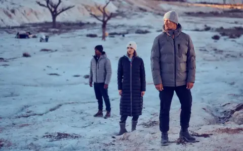 Drei Personen in Winterjacken stehen in einer verschneiten, kargen Landschaft. Die Frage "ab wann Winterjacke" stellt sich bei diesem Anblick von selbst.