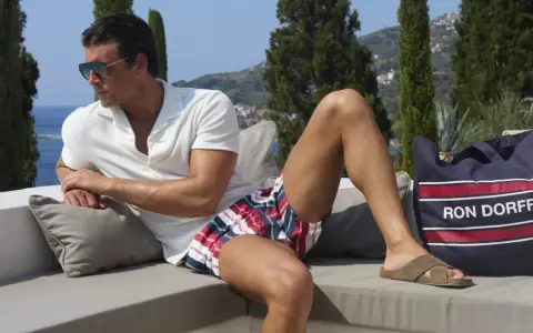 Ein Mann mit Sonnenbrille und gestreifter Badehose entspannt sich auf einer Couch. Neben ihm liegt eine Tasche mit der Aufschrift "RON DORFF".