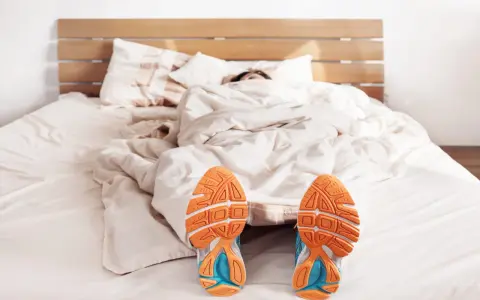 Füße mit Laufschuhen ragen aus einer Decke. Der Wecker klingelt, aber die Person träumt noch vom morgens joggen.