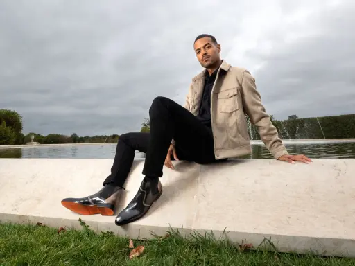 Mann in schwarzer Hose, beigem Sakko und stylischen Herren Boots, sitzt am Wasser.