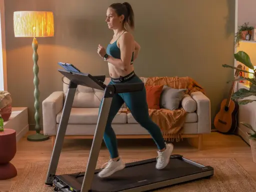 Frau macht Intervalltraining auf Laufband im Wohnzimmer. Sie trägt Sportkleidung und läuft mit Energie.