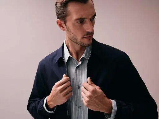 Ein Mann mit Bart und kurz geschnittenem Haar, der eine dunkelblaue Jacke über einem gestreiften Hemd trägt. Er fasst nach dem Reißverschluss, als würde er seine Kleidung für das Hemden waschen vorbereiten.