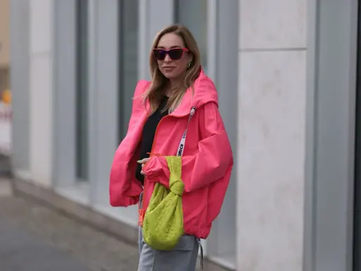 Stylische Frau in knallpinker Jacke und Sonnenbrille. Perfekt, um was anziehen bei 15 Grad. Dazu eine grüne Tasche.