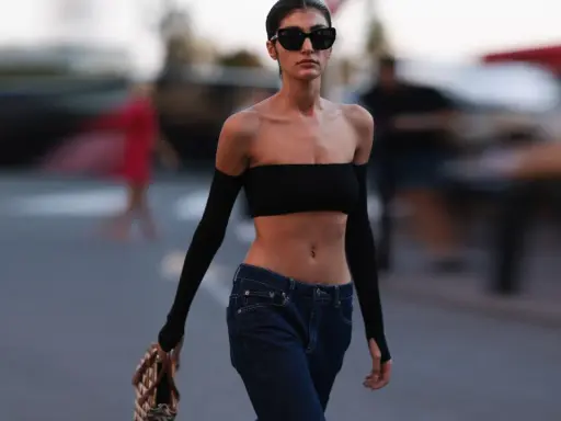 Frau in typischer 2000er Mode: schwarzer Bandeau-Top, tief sitzende Jeans und Armstulpen. Sie trägt Sonnenbrille und eine Tasche.