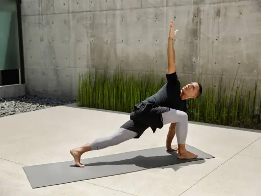 Mann in Yoga-Pose auf Matte vor Betonwand und grünen Pflanzen.
