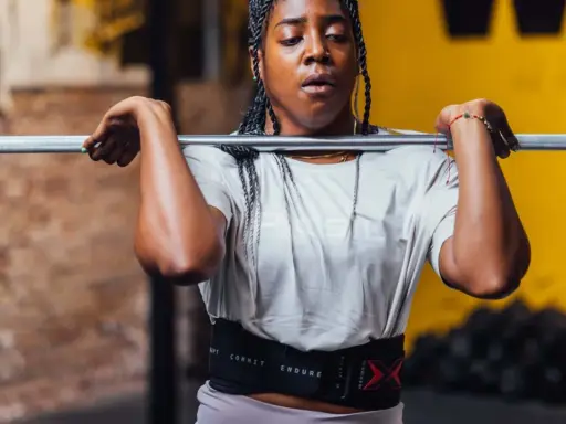 Frau mit geflochtenen Haaren hebt eine Hantel für Jumping Squats. Sie trägt einen Trainingsgürtel.
