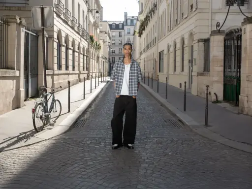 Ein Mann steht auf einer Kopfsteinpflasterstraße in Paris. Er trägt ein kariertes Hemd und weite Hosen. Ein Fahrrad lehnt an einem Laternenpfahl. Das Bild strahlt eine zeitlose Eleganz aus, die an reternity deutsch erinnert.